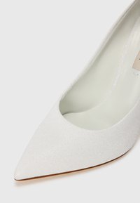 Hvide spidse pumps med tekstureret overdel, der har et subtilt glimt, og en lys inderside. Glat, tætsiddende design med minimale detaljer.