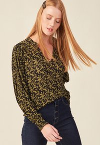 Blusa a maniche lunghe, con scollo a V, in stampa astratta verde e nera, caratterizzata da un tessuto testurizzato e dettagli di maniche gatherate. Abbinata a jeans a vita alta.