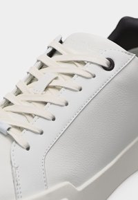 Λευκό χαμηλό αθλητικό παπούτσι με εκρού κορδόνια, δερμάτινα πάνελ με υφή και μαύρη εσωτερική επένδυση ορατή στη φτέρνα.