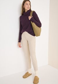 Maglione a collo alto a coste viola, pantaloni tapered crema, scarpe beige slip-on e una borsa marrone dalla texture morbida, in piedi davanti a uno sfondo neutro.