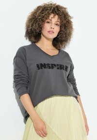 Felpa grigia scura con la parola "INSPIRE" in paillettes nere. Presenta un collo a V, maniche lunghe e una vestibilità comoda. Abbinata a una gonna fluida di un verde chiaro.