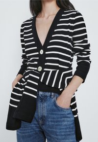 Czarny i biały kardigan w paski z głębokim dekoltem w serek, trzema guzikami z przodu oraz szczegółem w postaci paska w talii, zestawiony z niebieskimi dżinsami.