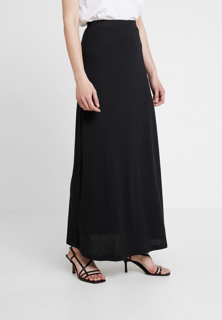 Femme portant une longue jupe noire, un haut blanc et des sandales noires à talons avec brides, debout devant un fond clair uni.