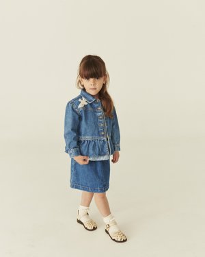 Junges Mädchen steht vor einem schlichten Hintergrund und trägt eine blaue Jeansjacke mit einem Schmetterlings-Aufnäher, dazu passenden Rock, weiße Socken und beige Sandalen.