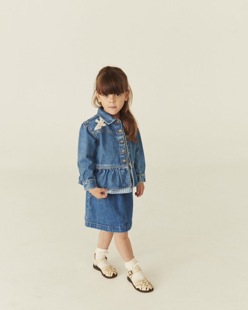 Noor tüdruk seisab sinist teksajopet, millel on liblikamärk, sobiva seelikuga, valgete sokkide ja beežide sandaalide seljas, ühevärvilise tausta ees.