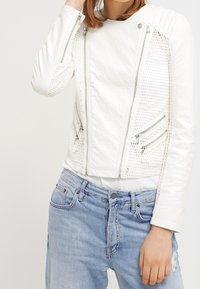 Vit kort bikerjacka med texturerat tyg, nätpaneler på sidorna, silverfärgad dragkedja och en figursydd design, kombinerad med ljusblå jeans.