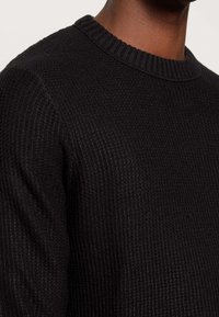 Gros plan sur une personne portant un pull en maille noire texturée avec un col rond côtelé et des manches longues.