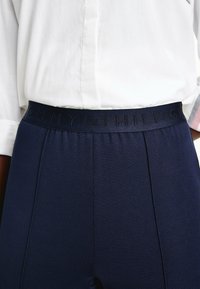 Laivansiniset housut, joissa on sileä kangas ja joustava vyötärö, jossa on kohokuvioitu tuotemerkin kirjain, yhdistettynä valkoiseen kauluspaitaan.