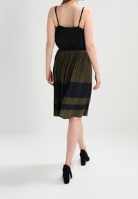 Débardeur noir associé à une jupe plissée longueur genou avec des rayures vertes et bleu marine. Le mannequin porte des chaussures à talons noires.