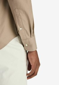 Ljusbrun långärmad skjorta med knappändar, som har en subtil tygstruktur och en böjd fåll. Handen vilar på vita byxor.