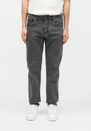 Osoba oblečená v vybledlých čiernych džínsach s rovnými nohavicami, bielych teniskách a bielej košeli zastrčenej dovnútra, stojaca pred jednoliatym pozadím.