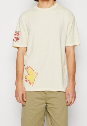 Mężczyzna w beżowej koszulce z grafiką czerwono-żółtego ptaka przy krawędzi i czerwonym napisem na lewej rękawie, w połączeniu z jasnobrązowymi spodniami.