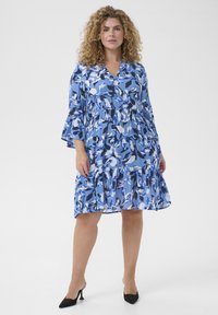 Blaues Blumen Kleid mit Glockenärmeln, einem V-Ausschnitt und einem gestuften Rock. Der Stoff zeigt verschiedene Blautöne mit weißen Akzenten.