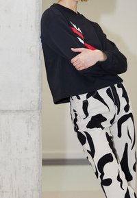 Personne appuyée contre un mur en béton portant un t-shirt noir à manches longues avec des rayures rouges et blanches et un pantalon à motif abstrait noir et blanc.