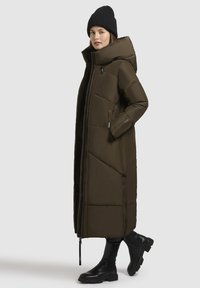 Dlhý, tmavozelený zateplený kabát s vysokým golierom, so zipsovými vreckami a rovnou siluetou. Nosený s čiernymi vysokými čižmami a čiapkou.