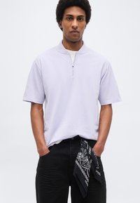 Νεαρός άνδρας με κοντά σγουρά μαλλιά, φορώντας ένα ανοιχτό μωβ κοντομάνικο πουκάμισο με φερμουάρ και μαύρο παντελόνι με μαύρο μπαντάνα κρεμασμένο από την τσέπη.