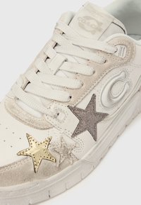 Balts Coach sporta apavu ar bēša zamšādas detaļām, daudzveidīgiem zvaigžņu motīviem zelta, sudraba un spīduma krāsās, un baltām auklām uz balta fona.