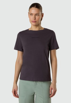 Donna con capelli castani raccolti indossa una maglietta North Sails viola scuro e pantaloni verde chiaro, guardando leggermente a sinistra.