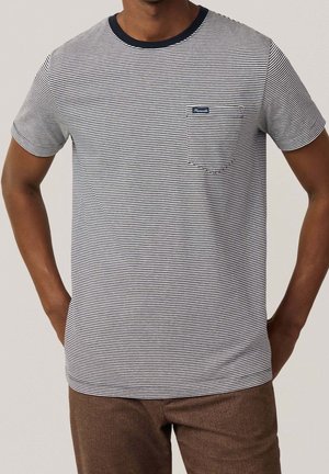 Man wearing a white and navy thin-striped short sleeve t-shirt with chest pocket and brown pants, hands resting on hips.