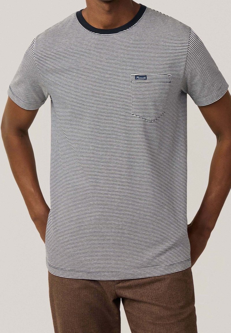 Homme portant un t-shirt à fines rayures blanches et bleu marine à manches courtes avec poche poitrine et pantalon marron, les mains posées sur les hanches.