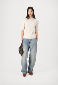 Cremfarbener, kurzärmeliger Strickpullover, kombiniert mit einer hellblauen, weiten Denim-Jeans. Das Model hält eine dunkle Ledertasche und trägt braune Schuhe.