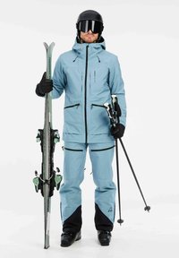 Ski-uitrusting met een lichtblauwe jas en broek met zwarte accenten. Accessoires omvatten groene ski's en zwarte skistokken. De outfit benadrukt functionaliteit.
