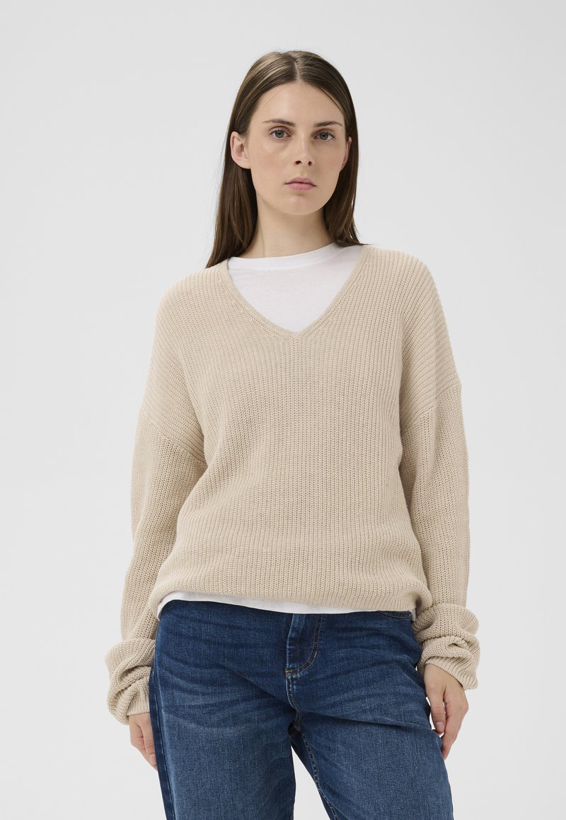 Femme aux cheveux bruns raides portant un pull en maille beige à col en V par-dessus une chemise blanche et un jean bleu, debout devant un fond uni.