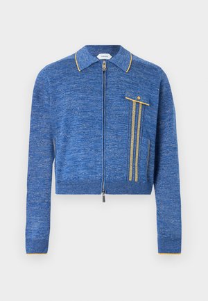 Mees kannab sinist lukuga kampsunit pruuni polo peal, mustad nahkpüksid ja mustad kingad, seisab ühetaolise helehalliga taustal.