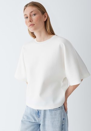 Jeune femme aux cheveux blonds lisses portant un haut blanc ample à manches courtes et un jean en denim bleu clair, sur un fond uni clair.