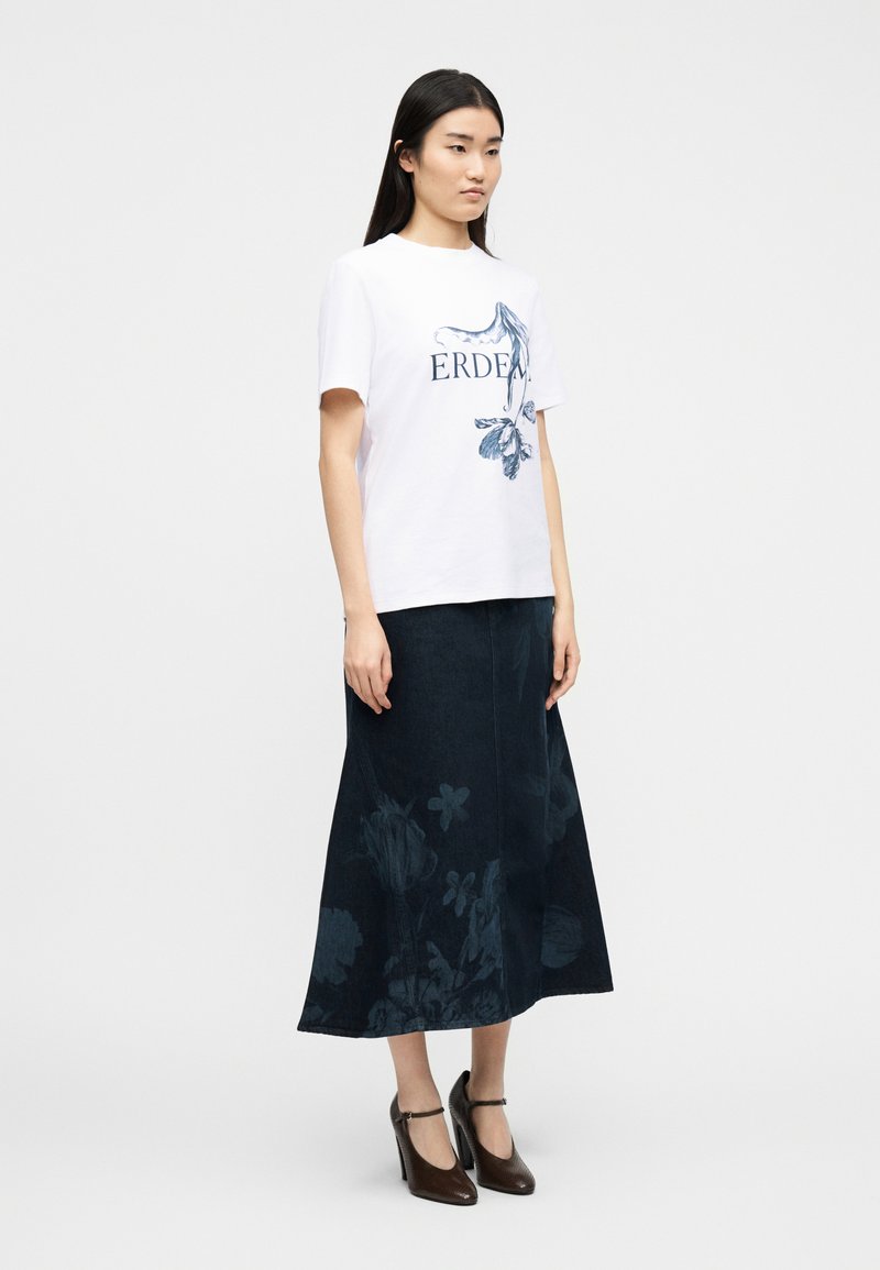 Vrouw draagt een wit T-shirt met grafisch ontwerp en de tekst "ERDEM", een donkerblauwe midi-rok met bloemenprint en bruine hoge hakken, staand tegen een eenvoudige achtergrond.