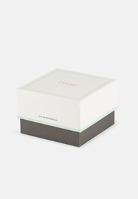 Boîte carrée avec un dessus blanc et un texte doré, présentant une bande vert menthe et un fond gris foncé texturé. Comprend un message "#CLUsetClub".
