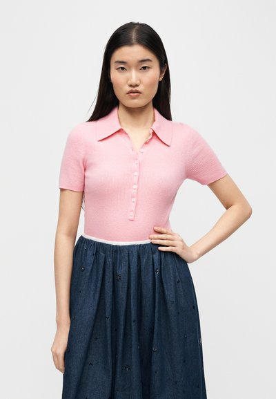 Νεαρή γυναίκα με μακριά σκούρα μαλλιά, φορώντας ένα ροζ πόλο πουλόβερ με κοντά μανίκια και κουμπιά, και μια ψηλόμεση σκούρο μπλε φούστα με μικρές διακοσμήσεις.