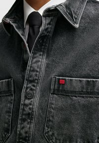 Giubbotto in denim nero con una texture a motivi. Presenta cuciture rosse, una chiusura con bottoni neri e un piccolo tag rosso con il marchio sulla tasca.