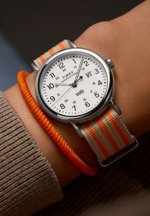 Montre Timex Weekender avec un boîtier rond en argent et un cadran blanc, comportant des index horaires noirs. Bracelet en nylon beige avec des rayures orange.