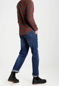 Czerwona i zielona koszula w kratę z długim rękawem o zaokrąglonym dole, zestawiona z niebieskimi dżinsami o tapered fit i czarnymi botkami Chelsea z brązową podeszwą.