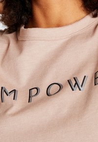 T-shirt in cotone rosa chiaro con la parola "EMPOWER" in lettere nere e grigie a contrasto. Collo rotondo, texture morbida.