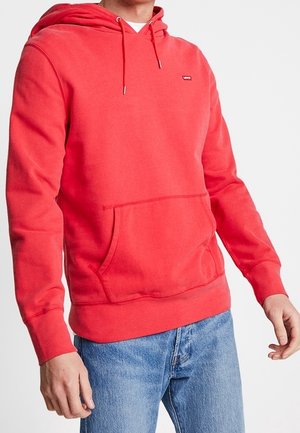 Mann trägt leuchtend roten Kapuzenpullover mit Fronttasche und hellblauen Jeans, steht vor einfachem Hintergrund.