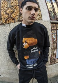 Sudadera negra con un gráfico de un oso de dibujo animado que lleva un gorro y el texto "GIANNI KAVANAGH" en la parte de atrás. El modelo lleva pantalones negros.