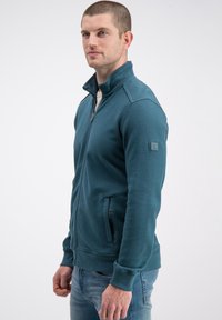 Morska zielona kurtka zapinana na zamek z wysokim kołnierzem, teksturowanymi panelami na rękawach i bocznymi kieszeniami. Casualowy design z prążkowanymi mankietami i dołem.