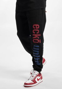 Black sweatpants with red and blue "eckō united" text along the legs, featuring a relaxed fit and elastic waistband. Paired with red and white sneakers.