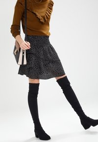 Schwarzer Rock mit Polka-Dots, schwingend mit Rüsche am Saum, kombiniert mit einem braunen Rippstrickpullover. Schwarze Stiefel aus Wildleder bis über das Knie und eine Umhängetasche.