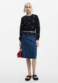 Felpa navy con ricami floreali, gonna midi in denim, cintura nera con borchie, borsa rossa e sandali in gomma neri.