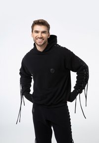 Czarna bluza z kapturem wykonana z miękkiego materiału, z ozdobnymi sznurówkami wzdłuż rękawów oraz okrągłą naszywką z logo na klatce piersiowej. Połączona z czarnymi spodniami.