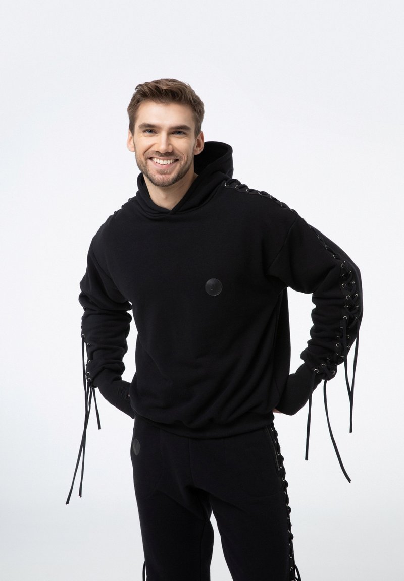 Czarna bluza z kapturem wykonana z miękkiego materiału, z ozdobnymi sznurówkami wzdłuż rękawów oraz okrągłą naszywką z logo na klatce piersiowej. Połączona z czarnymi spodniami.