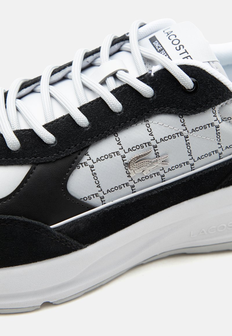 Sneaker Lacoste bianco e nero con lacci bianchi, motivo a ripetizione con testo Lacoste e logo del coccodrillo argentato sul pannello laterale.