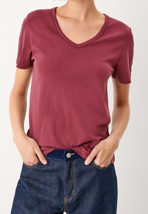 Vrouw draagt een bordeauxrode T-shirt met korte mouwen en V-hals, ingestopt in een donkerblauwe spijkerbroek, met handen in de zakken.