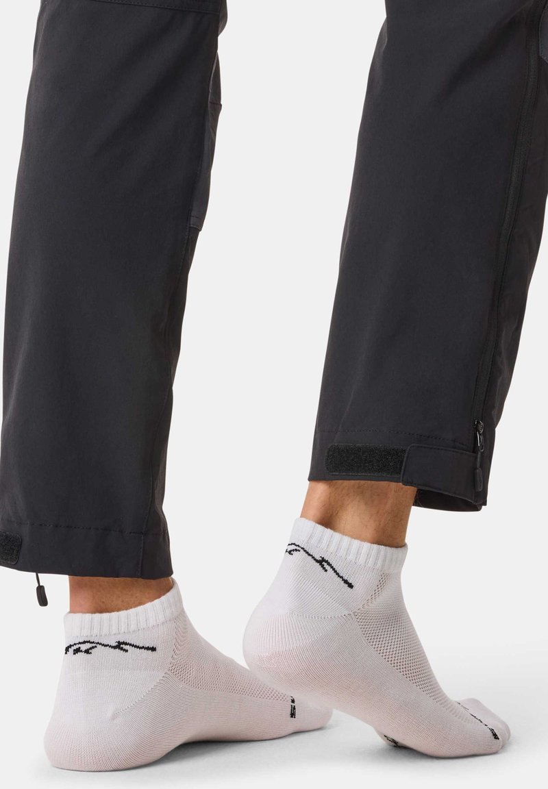 Deux pieds portant des chaussettes blanches aux chevilles avec un texte noir, un pied à plat et l’autre légèrement levé, pantalon noir coupé au-dessus des chevilles.