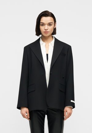 Žena s kratkom tamnom kosom, odjevena u preveliku crnu blejzerku, bijelu košulju s gumbima i crne hlače, stoji ispred jednobojne svijetle pozadine.