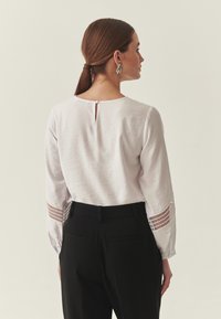 Blouse blanche à manches longues en tissu texturé, dotée d'une fermeture à boutons dans le dos et d'accents en ajouré sur les manches. Associée à un pantalon noir.