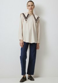 Lichtcrème blouse met een brede, puntige kraag met donkerbruine strepen, gecombineerd met donkerblauwe rechte jeans en zwarte instappers.